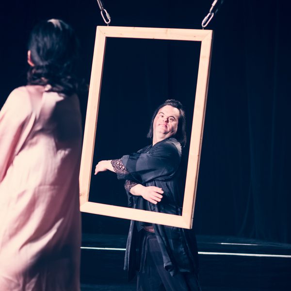 A man stands in front of a picture frame. A woman looks at him as if she were looking into a mirror.