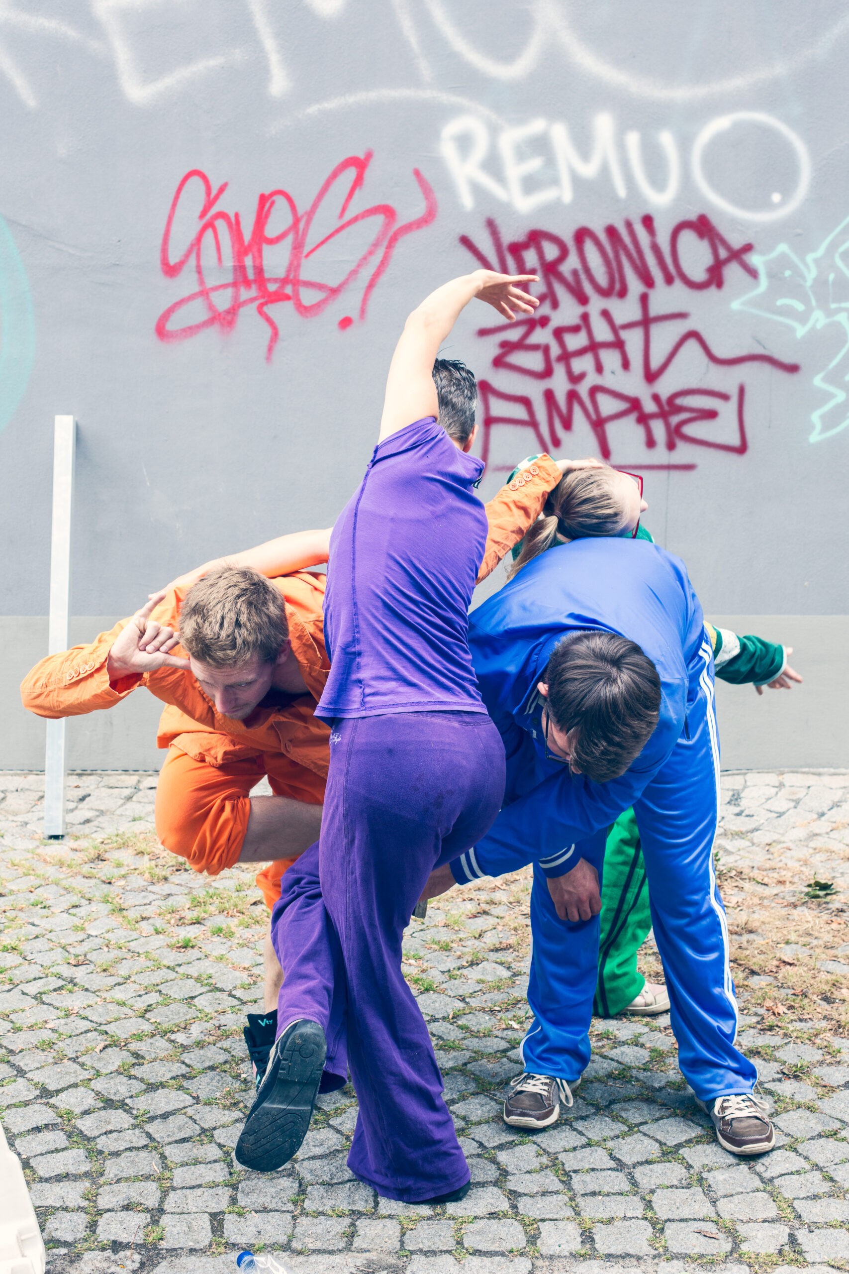 3 sehr bunt gekleidete Menschen vor einer Grafittiwand in einer Verknotung