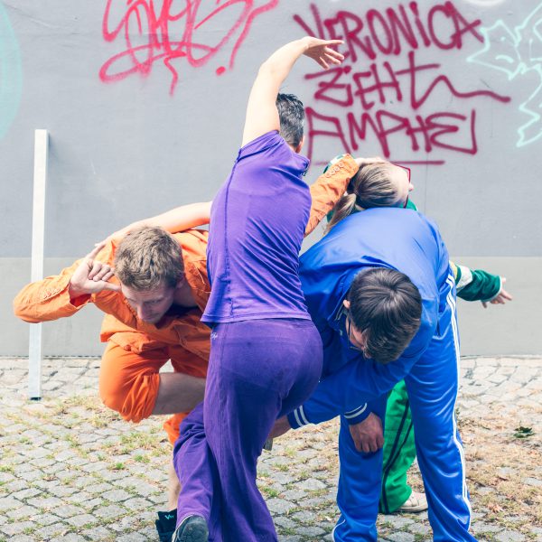 3 Tänzer:innen verknoten sich miteinander vor einer Grafitti-Wand