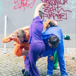 3 Tänzer:innen verknoten sich miteinander vor einer Grafitti-Wand