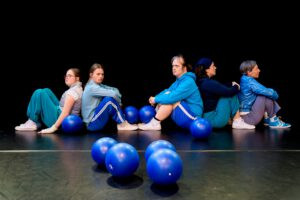 5 Tänzer:innen sitzen am Boden. Viele blaue Bälle liegen am Boden verstreut.