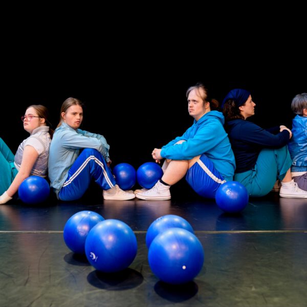 5 Tänzer:innen sitzen am Boden. Viele blaue Bälle liegen am Boden verstreut.