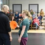 zwei Clowns von hinten aufgenommen auf der Bühne. Sie schauen sich an. Eine Clownin hält einen orangen Trichter in der Hand. Im Hintergrund sieht man Zuschauende auf der Tribüne