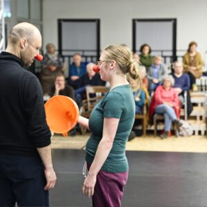 zwei Clowns von hinten aufgenommen auf der Bühne. Sie schauen sich an. Eine Clownin hält einen orangen Trichter in der Hand. Im Hintergrund sieht man Zuschauende auf der Tribüne