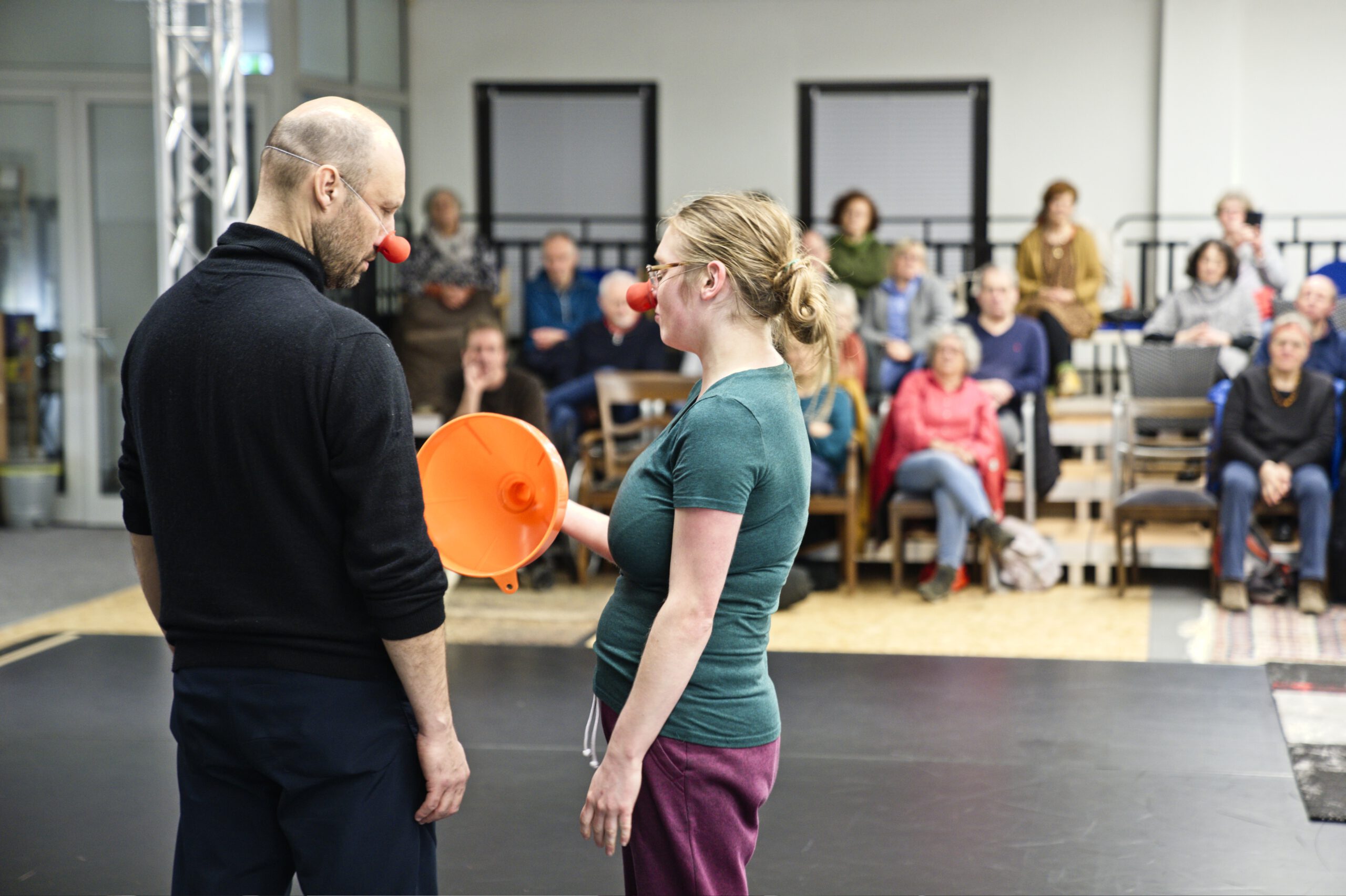 zwei Clowns von hinten aufgenommen auf der Bühne. Sie schauen sich an. Eine Clownin hält einen orangen Trichter in der Hand. Im Hintergrund sieht man Zuschauende auf der Tribüne