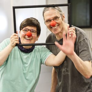 Zwei Clown:innen halten einen Bilderrahmen und schauen gemeinsam hindurch