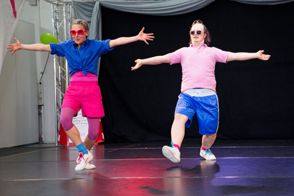 zwei Tänzer:innen in Rosa und Blau gekleidet, mit Sonnenbrillen. Sie haben die Arme weit, wie für eine Umarmung geöffnet und schauen ins Publikum