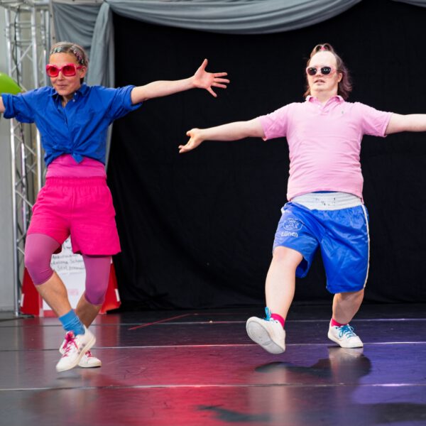 zwei Tänzer:innen in Rosa und Blau gekleidet, mit Sonnenbrillen. Sie haben die Arme weit, wie für eine Umarmung geöffnet und schauen ins Publikum