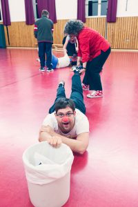 A smiling man with a red nose lies on the ground holding a bucket. A person pulls him back by the leg.