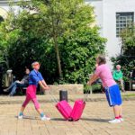 Zwei Tänzeri:nnen stehen sich mit aneinandergeschobenen Koffern gegenüber. Sie tragen Pink und Blau