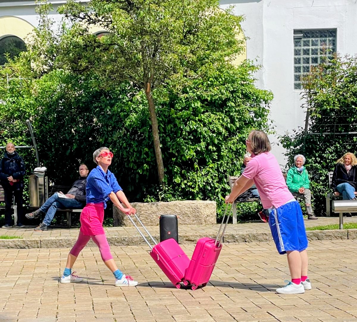 Zwei Tänzeri:nnen stehen sich mit aneinandergeschobenen Koffern gegenüber. Sie tragen Pink und Blau