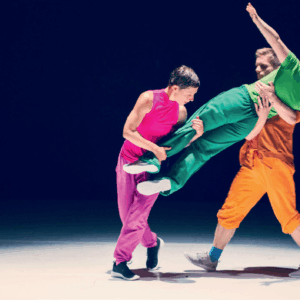 3 Tänzerinnen. eine in pink, eine in Grün, einer in Orange. Pink und Orange heben die Tänzerin in Grün.