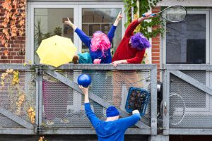 4 Personen mit Bällen, Schirmen, Kisten und bunten Perücken bilden eine Installation am Geländer.