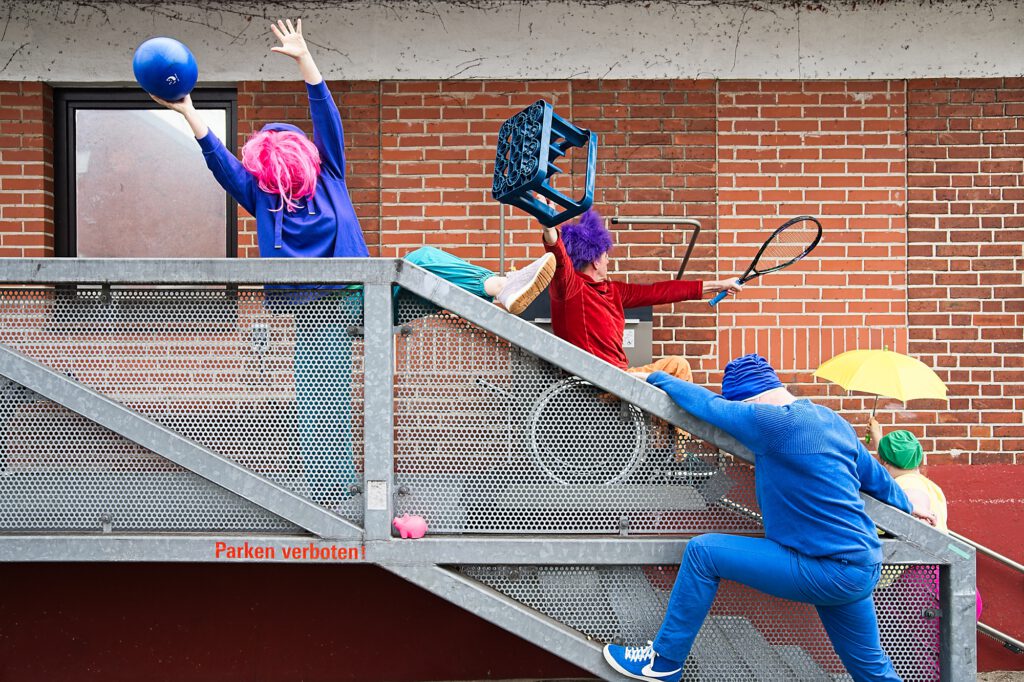 Eine Reihe bunter Gestalten mit Perücke, Kiste, Ballon, Tennisschläger posiert in auf der Treppe vor einem Geländer