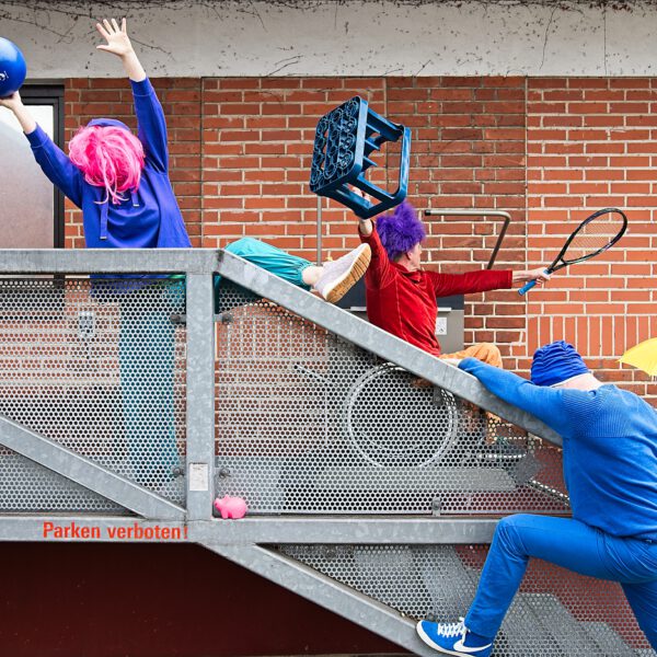 Eine Reihe bunter Gestalten mit Perücke, Kiste, Ballon, Tennisschläger posiert in auf der Treppe vor einem Geländer