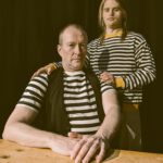 zwei Männer am Tisch, beide tragen gestreifte Seemannspullis. Einer sitzt, einer steht hinter dem Sitzenden und hat die Hände auf seine Schultern gelegt. Beide schauen in die Kamera