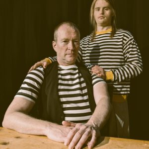 zwei Männer am Tisch, beide tragen gestreifte Seemannspullis. Einer sitzt, einer steht hinter dem Sitzenden und hat die Hände auf seine Schultern gelegt. Beide schauen in die Kamera