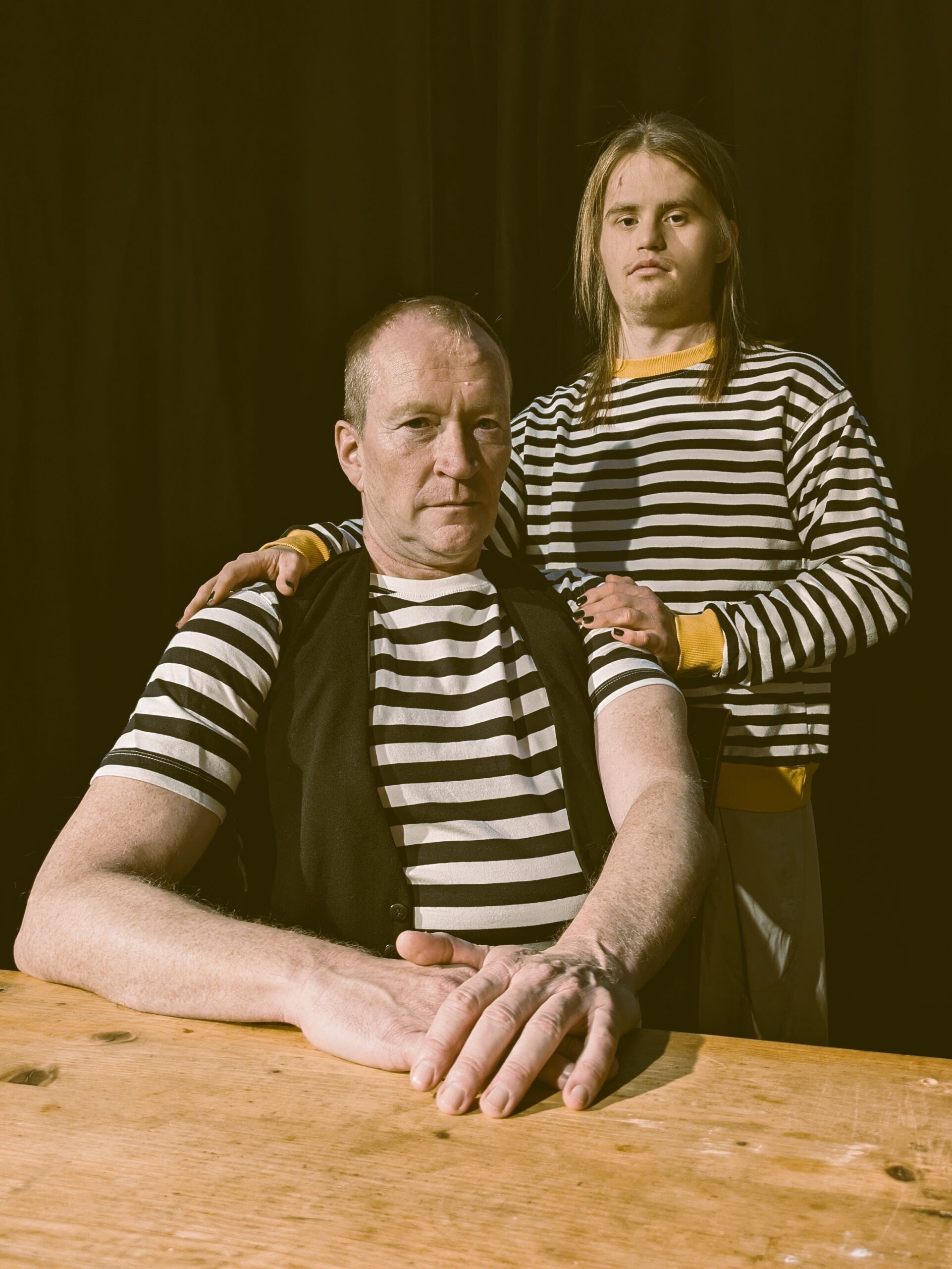 zwei Männer am Tisch, beide tragen gestreifte Seemannspullis. Einer sitzt, einer steht hinter dem Sitzenden und hat die Hände auf seine Schultern gelegt. Beide schauen in die Kamera