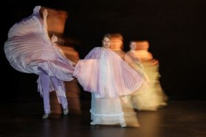 3 Tänzer:innen in langen, plissierten Umhängen in Bewegung. Das Bild ist lang belichtet, weswegen die Umrisse verschwommen und in Bewegung zu sein scheinen.