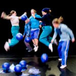 5 in die Luft springende Tänzer:innen. Am Boden liegen blaue Bälle