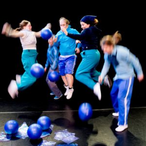 5 in die Luft springende Tänzer:innen. Am Boden liegen blaue Bälle