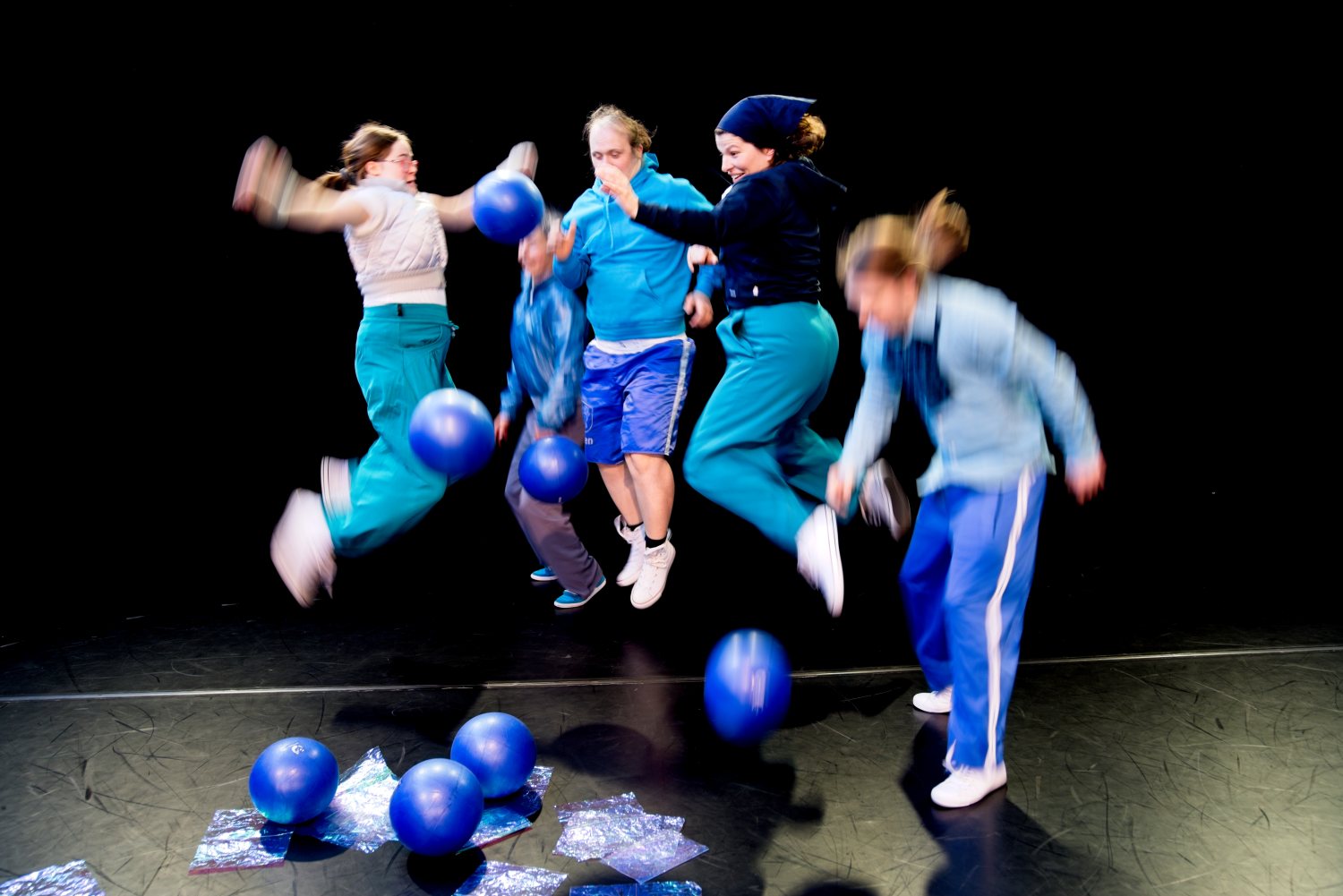 5 in die Luft springende Tänzer:innen. Am Boden liegen blaue Bälle
