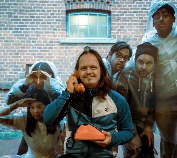 Adrian mit Telefon vor einer Glasscheibe. Dahinter drücken sich Menschen die Nasen platt