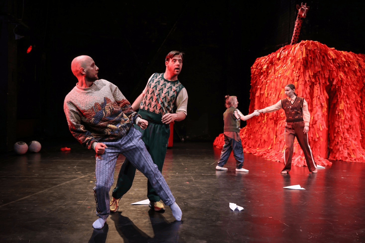 Zwei Männer tanzen im Vordergrund, zwei Performerinnen reichen sich im Hintergrund vor einem roten Lametta-Zelt die Hände