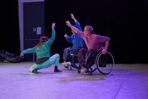 3 bunt gekleidete Tänzer:innen auf einer in lila Licht getauchten Bühne. 2 Tanzende im Rollstuhl erstellen eine Form mit den Armen, eine 3 Tänzerin hockt am Boden und nimmt die Bewegung der Arme auf